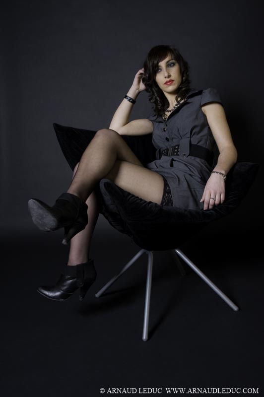 femme brune cheveux longs, maquillée, veste à épaulettes grises, bas et bottines noirs, assise jambes croisées sur un fauteuil en velours noir devant un fond  anthracite