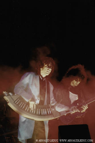 Jean-Michel Jarre dans un halo de fumigènes en concert au stade véllodrome de marseille, en veste longue bleu ciel,  jouant sur une orgue électrique semi circulaire portée en bandouillère, un musicien bassiste en blouson de cuir noir à sa droite