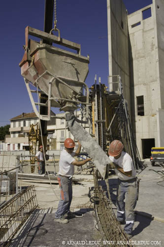 2 ouvriers sur un chantier de construction guidant l'écoulement du béton venant d'une benne suspendue à une grue, sur le feraillage des fondations d'un mur, ciel bleu en arrière plan