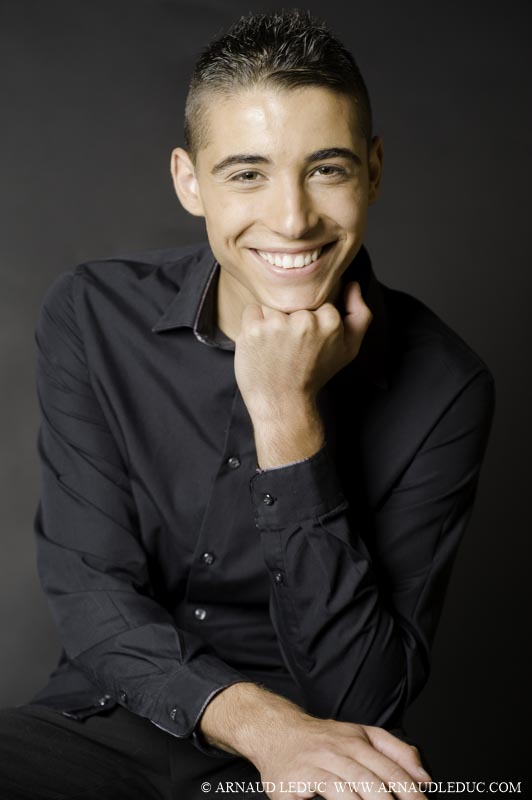 Jeune homme brun, cheveux cours, en chemise noire, souriant, son visage posé sur son poing gauche fermé