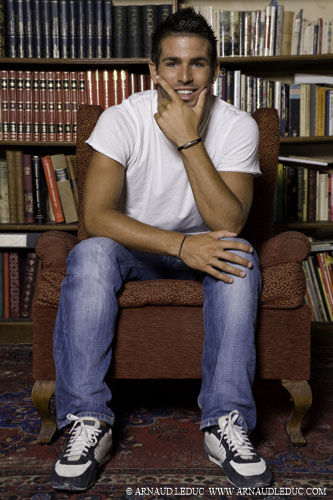 jeune homme brun cheveux courts en jean tshirt blanc et baskets assis de face dans un fauteuil en velours rouge devant une bibliothèque souriant, son visage posé sur sa main gauche
