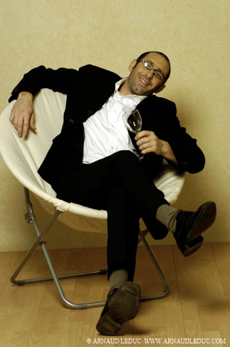 l'acteur Emmanuel Gueret en costume noir et chemise blanche assis dans un fauteuil pliant blanc, un verre a la main