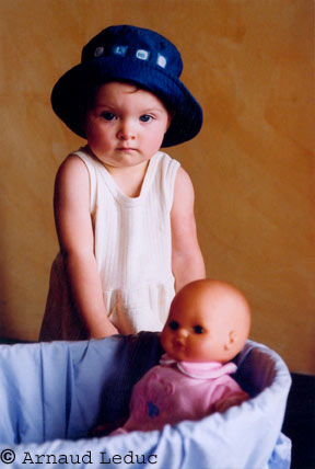 petite fille en robe creme et chapeau bleu devant une poupee dans un couffin bleu
